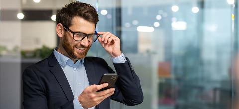 Successful financier investor works inside office at work, businessman in business suit uses telephone near window, man smiles and reads good news online from smartphone.