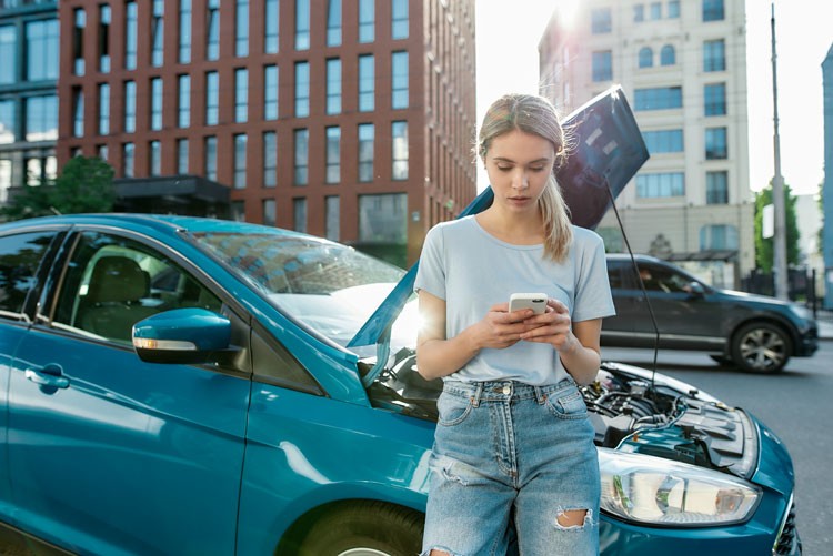 Attractive young woman holding phone, calling emergency car service while standing, leaning on her broken car with open hood on the city street