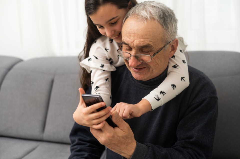 senior grandfather learning to using mobile phone under guidance of pretty young granddaughter sitting on sofa at home