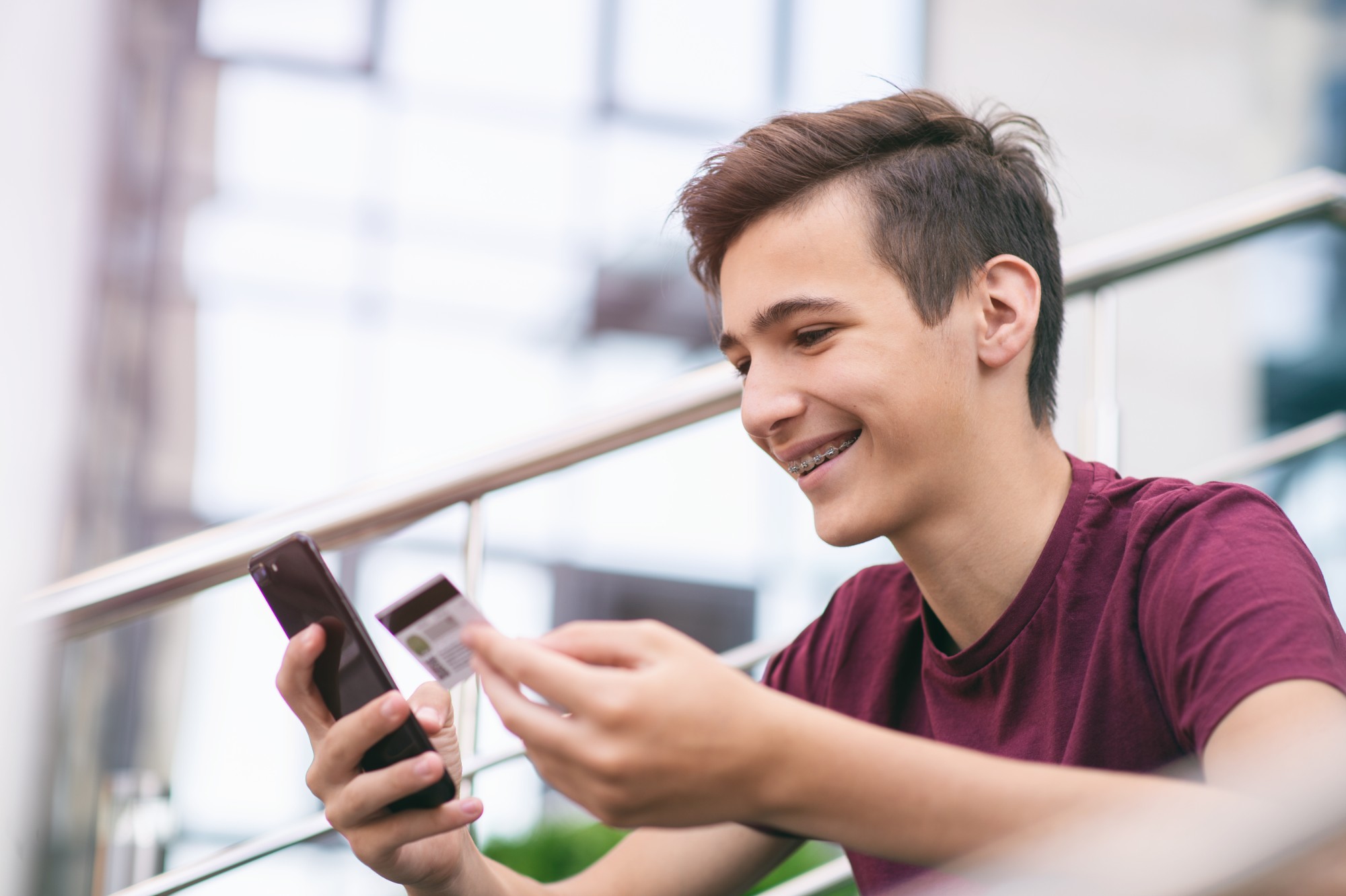 Teenage boy with a credit card and mobile phone makes purchasing outdoors. Happy young man is using smartphone and bank card for online shopping. Handsome smiling guy holds bank card and cell phone