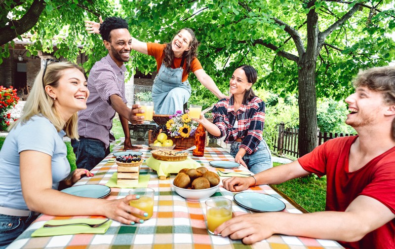 Young men and women toasting healthy orange fruit juice at farm house patio picnic - Life style concept with happy friends having fun together on afternoon relax time - Bright vivid filter