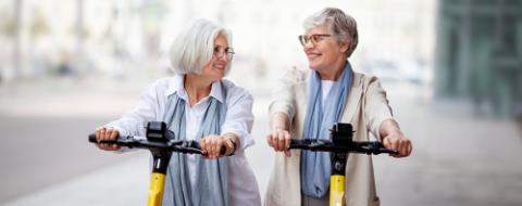 Mature joyful gray haired women friends ride electric scooters in the city, happy smiling, enjoying a fun time together