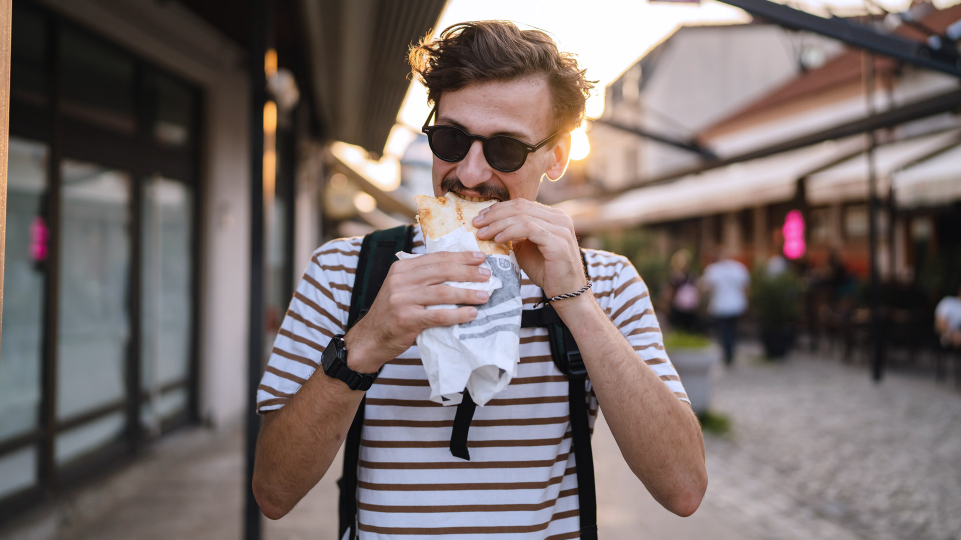Een jonge man staat aan een station. De man eet een broodje en heeft net een (terug)betaling via zijn smartphone te hebben verstuurd of ontvangen. Het beeld straalt gemak en snelheid uit — alsof hij onderweg snel iets regelt zonder cash of bankkaart.